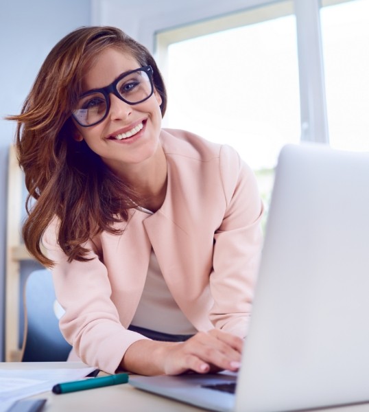 Smiling woman in glasses and pale pink jacket sitting at desk with laptop